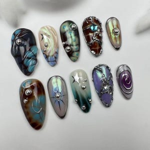 May include: Ten different nail designs featuring various colors, patterns, and embellishments. The designs include a swirl, a star, a galaxy, a spiderweb, and a seashell.