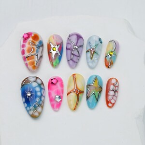 May include: A set of ten press-on nails featuring various colorful designs, including abstract patterns, gradients, and marble effects. Several nails are decorated with metallic silver star-shaped accents, small rhinestones, and intricate silver line work.