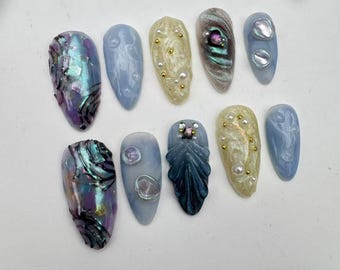 May include: Set of ten false nails with various designs. The nails are a light blue color with different embellishments, including iridescent shells, pearls, and glitter.
