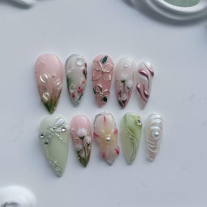 May include: A set of ten almond-shaped artificial nails. The nails feature various designs, including floral patterns, dragonfly accents, and pearl embellishments. Colors include pink, white, green, and silver, with metallic and 3D elements.