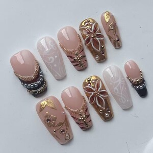 May include: A collection of artificial nails in various designs. Some nails are nude with gold chains and crosses, others feature gold floral designs, and some have white lace patterns. The nails are adorned with pearls and gems.