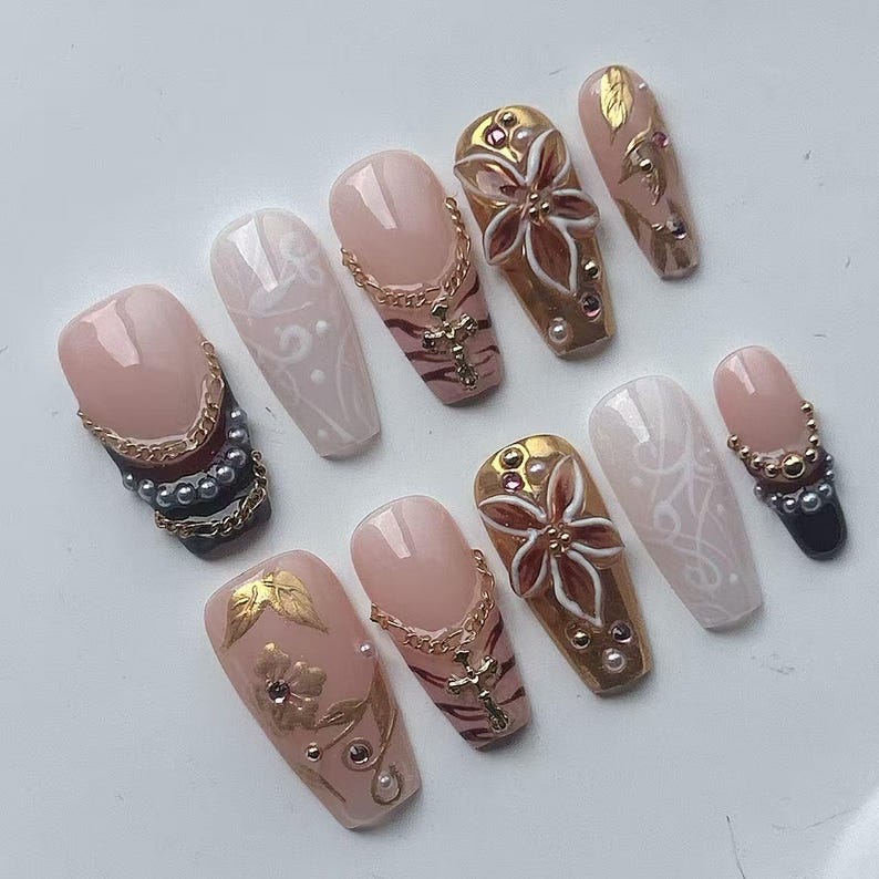 May include: A collection of artificial nails in various designs. Some nails are nude with gold chains and crosses, others feature gold floral designs, and some have white lace patterns. The nails are adorned with pearls and gems.