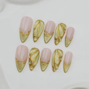 May include: A set of ten almond-shaped false nails. The nails feature a pale pink base with gold accents. Some nails have a gold butterfly wing design, while others have gold trim along the tips.