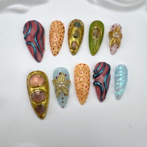 Puede incluir: Diez uñas artificiales con diversos diseños. Las uñas presentan colores como oro, rosa, azul y verde. Algunas uñas tienen espirales, puntos y adornos florales. Otras tienen un aspecto texturizado e iridiscente.