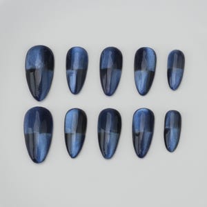 May include: Set of ten oval-shaped false nails in varying sizes. Each nail is a deep blue with a shimmering, cat-eye effect. The nails are arranged on a white surface, showcasing their glossy finish and color variations.