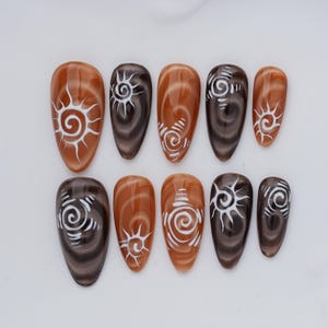 Puede incluir: Un juego de diez uñas postizas en forma de almendra con efecto ojo de gato en tonos alternos de marrón ámbar cálido y marrón espresso profundo. Cada uña está decorada con un diseño de sol en espiral blanco pintado a mano con líneas radiantes.