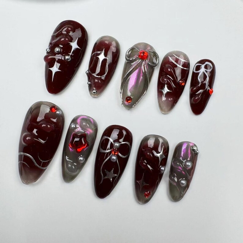 May include: A set of ten artificial nails in various shapes and designs. The nails feature a dark red base with silver accents, including stars, bows, and swirls. Some nails have iridescent and pearl embellishments.