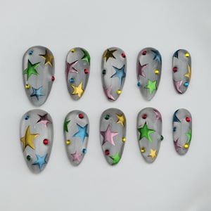 Puede incluir: Juego de diez uñas postizas transparentes de forma ovalada. Cada uña presenta un diseño de estrella colorida en tonos amarillo, verde, azul y rosa, con pequeños acentos de gemas coloridas. Las uñas están dispuestas sobre una superficie blanca.
