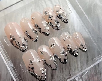 May include: A set of ten nude-colored false nails with silver glitter and intricate silver designs. The nails are displayed in a clear plastic box.