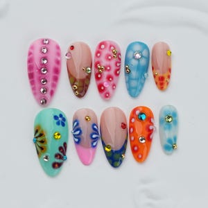 May include: A set of ten almond-shaped press-on nails featuring diverse designs including pink with rhinestones, blue and orange floral patterns, polka dots, and multi-colored gems. Each nail displays unique artistic details and embellishments.
