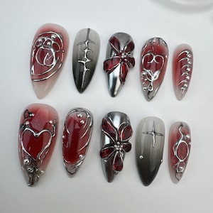 May include: A set of ten artificial nails with a variety of designs. The nails feature a combination of red, silver, and black colors. Some nails have a gradient effect, while others showcase intricate silver embellishments and floral or heart-shaped designs.