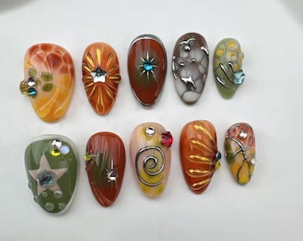 Könnte beinhalten: Zehn dekorative Nagelspitzen mit verschiedenen Designs, darunter Spiralen, Sterne und Blumen. Die Spitzen sind in Orange-, Grün- und Brauntönen lackiert.