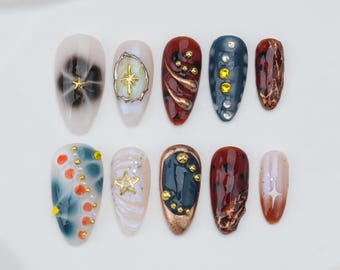 Könnte beinhalten: Eine Sammlung von zehn künstlichen Nägeln mit verschiedenen Designs. Die Nägel weisen eine Reihe von Farben auf, darunter Weiß, Rot, Blau und Gold. Einige Nägel haben Stern- und Edelsteinverzierungen, während andere abstrakte Muster und Texturen aufweisen.