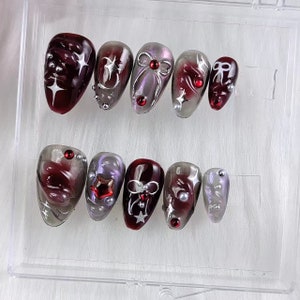 May include: A set of ten artificial nails in a clear plastic case. The nails feature a dark red and silver color scheme with various designs, including stars, bows, and embellishments. Some nails have a marbled effect.