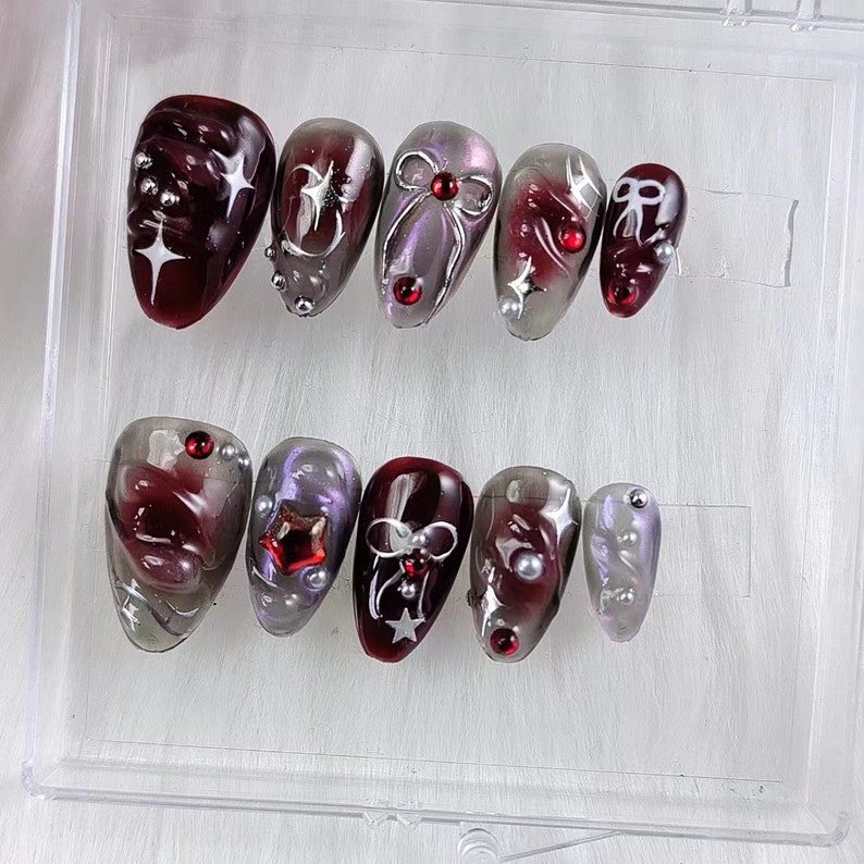 May include: A set of ten artificial nails in a clear plastic case. The nails feature a dark red and silver color scheme with various designs, including stars, bows, and embellishments. Some nails have a marbled effect.