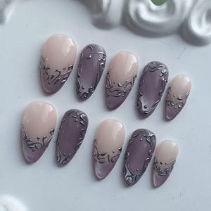 May include: Set of ten almond-shaped false nails in a variety of sizes. The nails are painted in a combination of mauve and pale pink, with silver filigree designs. The nails are arranged on a white surface.