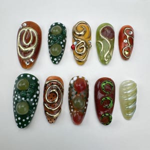 May include: A collection of ten uniquely designed artificial nails. Each nail features a different color scheme, including shades of green, orange, red, and yellow, with gold accents and decorative embellishments. Some nails have a swirl design, while others have dots or gems.