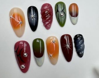 Könnte beinhalten: Set mit zehn falschen Nägeln mit verschiedenen Designs. Die Nägel sind in verschiedenen Farben gehalten, darunter Orange, Rot, Grün und Schwarz. Einige Nägel haben ein Spiralmuster, während andere ein Spinnennetzmuster haben. Alle Nägel haben eine klare Basis und sind mit silbernen Strasssteinen verziert.