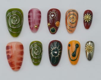 Könnte beinhalten: Ein Set aus zehn künstlichen Nägeln in verschiedenen Designs und Farben. Die Nägel zeigen Wirbel, Sonnenstrahlen und abstrakte Muster in Grün, Rot, Orange und Gold. Einige Nägel haben goldene Verzierungen, andere einen Marmoreffekt.