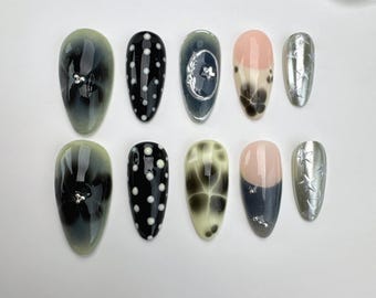 Pode incluir: Um conjunto de dez unhas artificiais com vários designs. As unhas apresentam padrões, incluindo bolinhas pretas e brancas, uma lua crescente e um design de estrela prateada. Algumas unhas têm um efeito gradiente ou são adornadas com pequenos strass.