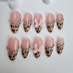 Può includere: Set di dieci unghie finte con un design a stampa leopardata. Ogni unghia ha una base trasparente con un bordo di strass argentati. Due unghie presentano un fiocco rosa.