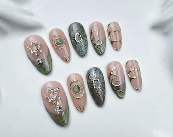 May include: Set of ten false nails with a light pink base and green glitter. Some nails have gold accents, including a sun, a crescent moon, and a star.