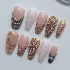 May include: A set of ten artificial nails in various designs. The nails feature a range of colors, including pink, gold, and white, with embellishments such as gold chains, crosses, pearls, and floral patterns. Some nails have a matte finish, while others are glossy.