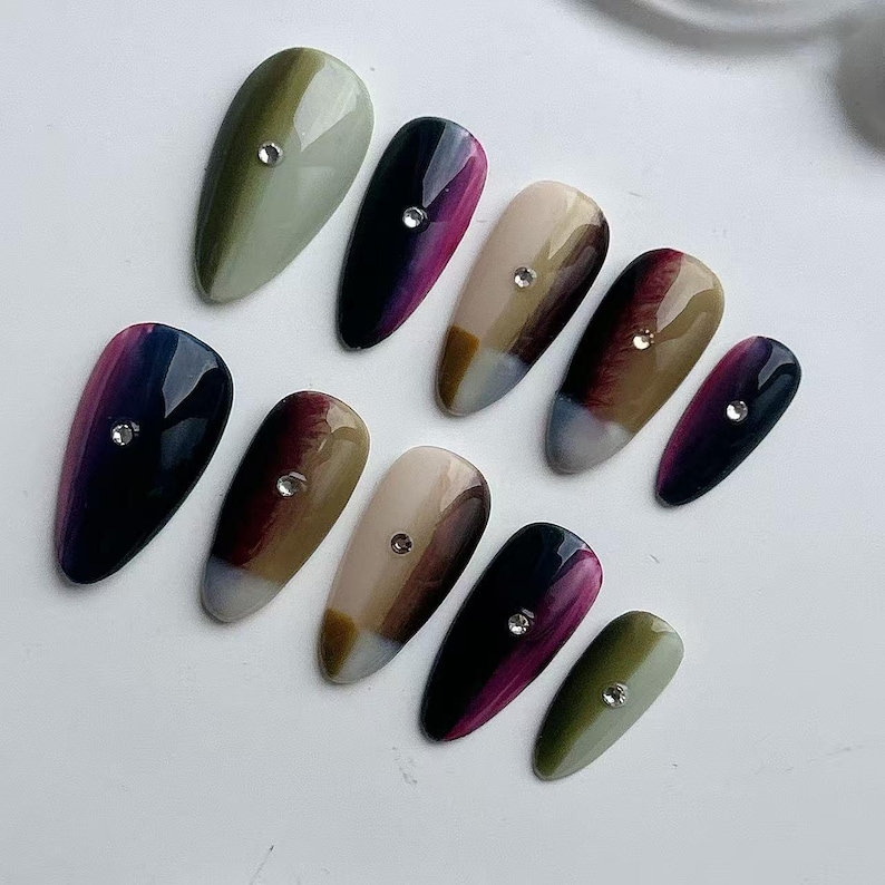 Puede incluir: Una colecci&oacute;n de diez u&ntilde;as artificiales de forma ovalada. Cada u&ntilde;a presenta un degradado de color &uacute;nico, que va del verde oliva al p&uacute;rpura oscuro, con un peque&ntilde;o brillante transparente cerca de la base. Las u&ntilde;as est&aacute;n dise&ntilde;adas para uso cosm&eacute;tico.