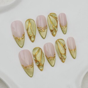 May include: A set of ten almond-shaped false nails. Each nail features a pale pink base with a gold design. The designs include gold accents along the tips and intricate patterns resembling leaves or wings. The nails are arranged on a white surface.