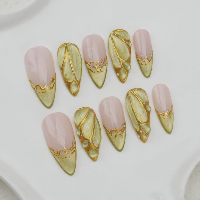 May include: A set of ten almond-shaped false nails. Each nail features a pale pink base with a gold design. The designs include gold accents along the tips and intricate patterns resembling leaves or wings. The nails are arranged on a white surface.