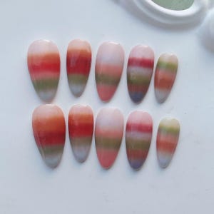 Peut inclure: Un ensemble de dix faux ongles ovales. Chaque ongle présente un dégradé avec des rayures horizontales orange, rouge, vert et violet, se fondant en une pointe blanche. Les ongles sont disposés sur une surface blanche.