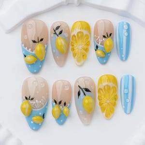 May include: A set of ten press-on nails featuring a lemon theme. The designs include yellow 3D lemon fruit, sliced lemon segments, light blue and white stripes, and polka dots, accented with clear raised water droplet details on a nude base.