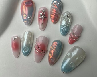 May include: A set of ten pastel-colored fake nails with various designs. The nails feature iridescent finishes, pearl accents, and silver chains.