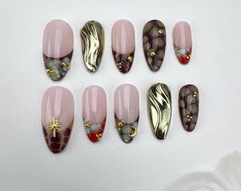 Könnte beinhalten: Ein Set aus zehn künstlichen Nägeln in verschiedenen Designs. Die Nägel haben eine hellrosa Basis mit goldenen, roten und braunen Akzenten. Einige Nägel haben ein goldenes Wirbeldesign, andere goldene Stern- oder Monddetails.