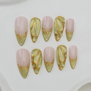 May include: A set of ten almond-shaped false nails. The nails feature a light pink base with gold accents. Some nails have a gold design at the tip, while others have a gold and green butterfly wing design.