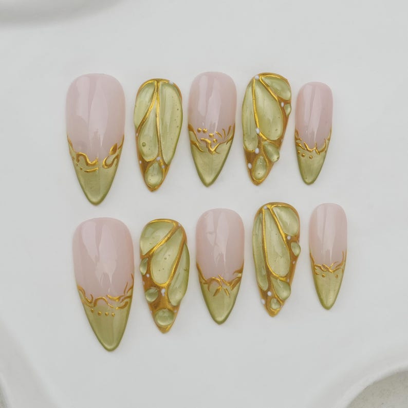 May include: A set of ten almond-shaped false nails. The nails feature a light pink base with gold accents. Some nails have a gold design at the tip, while others have a gold and green butterfly wing design.