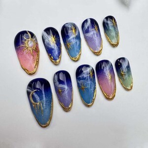 May include: Set of ten false nails with a celestial theme. The nails are painted in shades of blue, pink, and green with gold accents. Each nail features a different design, including moons, stars, and clouds.