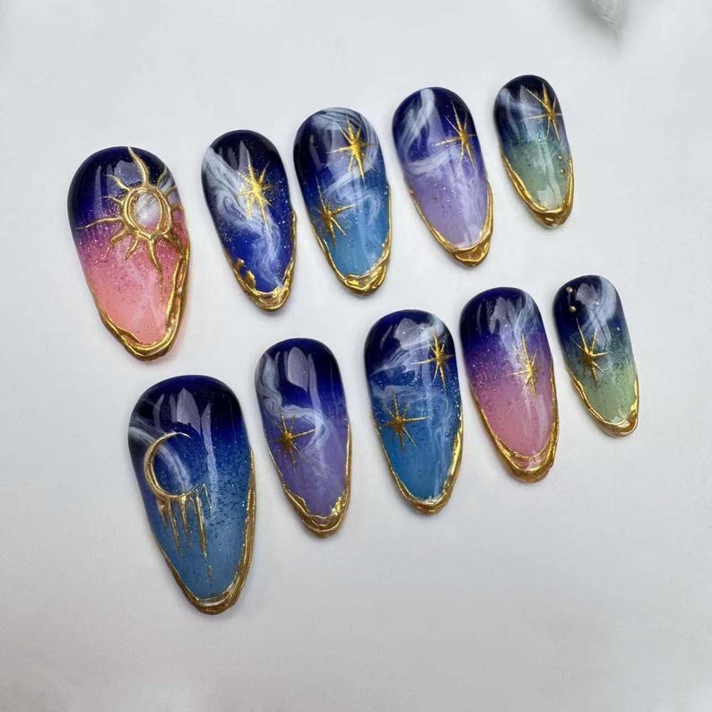 May include: Set of ten false nails with a celestial theme. The nails are painted in shades of blue, pink, and green with gold accents. Each nail features a different design, including moons, stars, and clouds.
