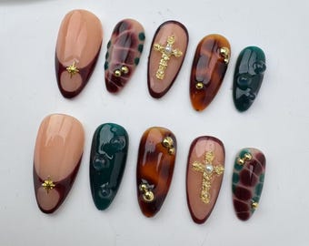 Könnte beinhalten: Satz von zehn falschen Nägeln mit verschiedenen Designs. Einige Nägel haben ein goldenes Kreuz mit einem Perlenakzent, andere ein Schildpattmuster und einige ein grünes und burgunderfarbenes Wirbelmuster.