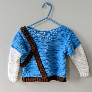 Könnte beinhalten: Ein blauer Häkelpullover mit braunen Trägern und weißen Ärmeln. Der Pullover hat einen V-Ausschnitt und ist mit weißen Nähten verziert.