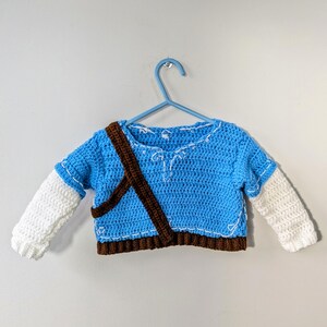 Puede incluir: Un suéter de crochet azul con mangas blancas y una correa de crochet marrón. El suéter tiene un diseño de crochet blanco en la parte delantera.