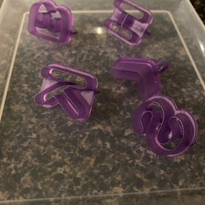 Op de afbeelding: Vijf paarse plastic koekjesvormen in een doorzichtige plastic container. De vormen zijn in de vorm van een hart, een letter H, een letter R, een letter S en een wolk.