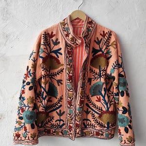 Puede incluir: Chaqueta color melocotón con intrincados bordados florales en tonos azules, verdes y marrones. La chaqueta tiene cuello mandarín y forro naranja a rayas. El bordado es detallado y cubre el frente, las mangas y el cuello.