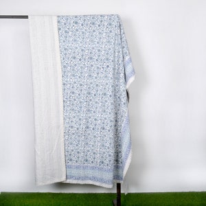May include: A white and blue kantha quilt with a floral pattern. The quilt is folded and hanging on a rack.