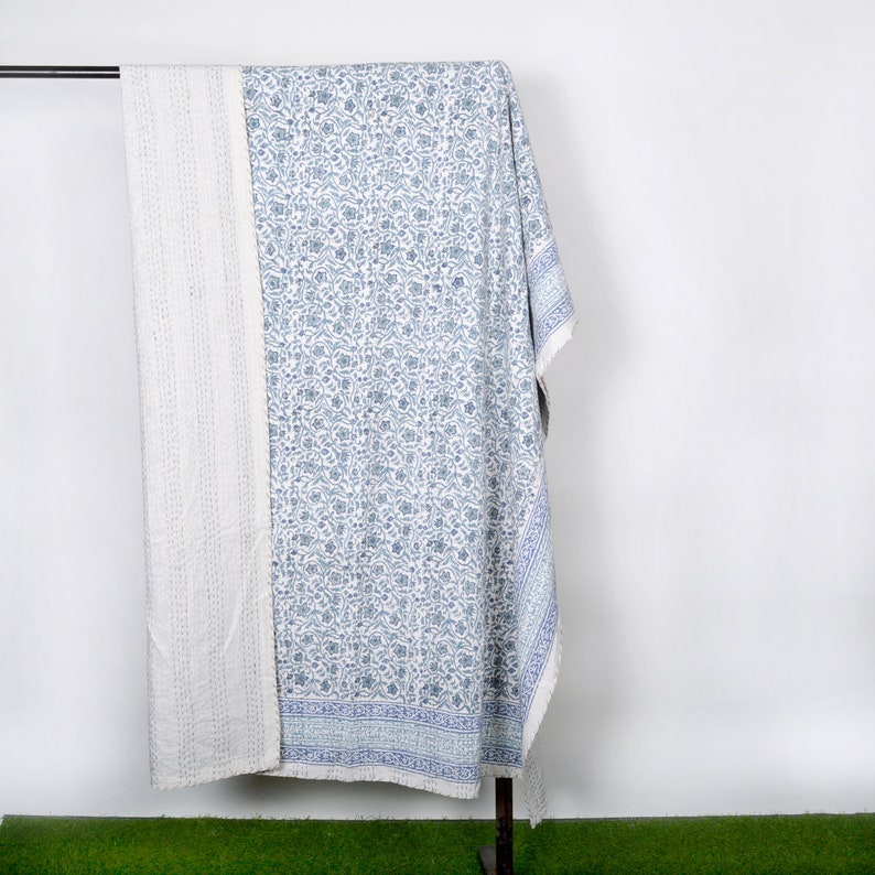 May include: A white and blue kantha quilt with a floral pattern. The quilt is folded and hanging on a rack.