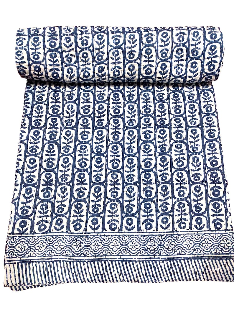 K&ouml;nnte beinhalten: Eine wei&szlig;e und blaue Kantha-Steppdecke mit einem floralen Muster. Die Steppdecke ist in der H&auml;lfte gefaltet und hat einen dekorativen Rand.
