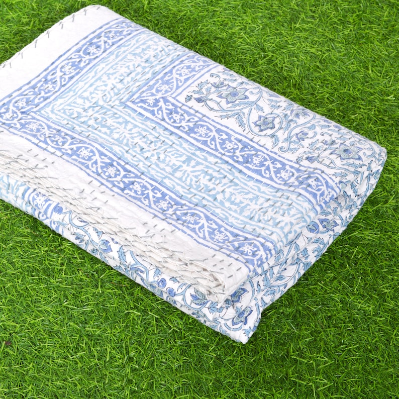 May include: A white cotton quilt with a blue and white floral pattern. The quilt is folded and lying on a green grassy surface.