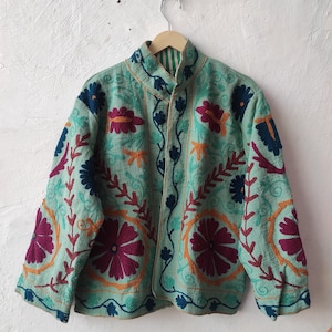 Puede incluir: Chaqueta turquesa con intrincados bordados florales en tonos morados, naranjas y azules. La chaqueta tiene un cuello alto y se muestra en una percha de madera. El bordado crea un aspecto texturizado y decorativo.