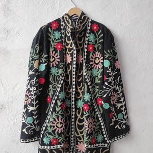 Pode incluir: Casaco preto bordado com padrões florais coloridos em vermelho, rosa, azul e amarelo. O casaco está pendurado num cabide de madeira contra uma parede branca. O bordado é detalhado e cobre a frente e as mangas.