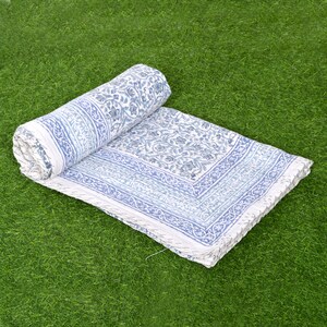 May include: A white cotton quilt with a blue floral pattern. The quilt is rolled up and lying on a green grassy surface.